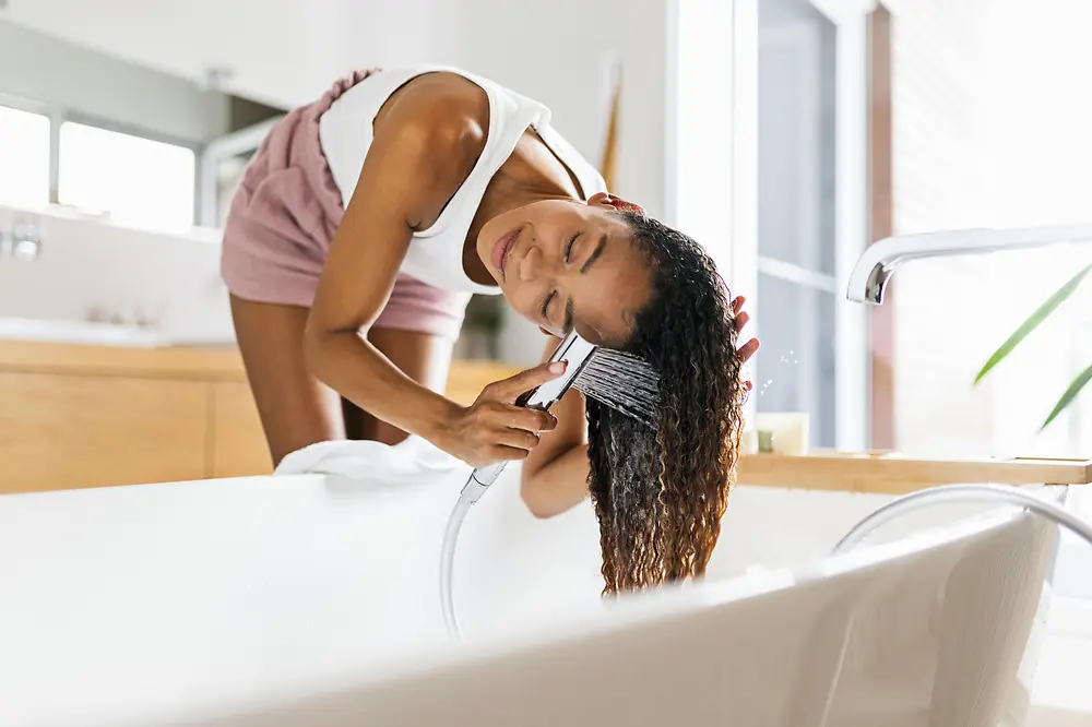 Žena si nad vaňou v kúpeľni umýva dlhé hnedé vlasy
