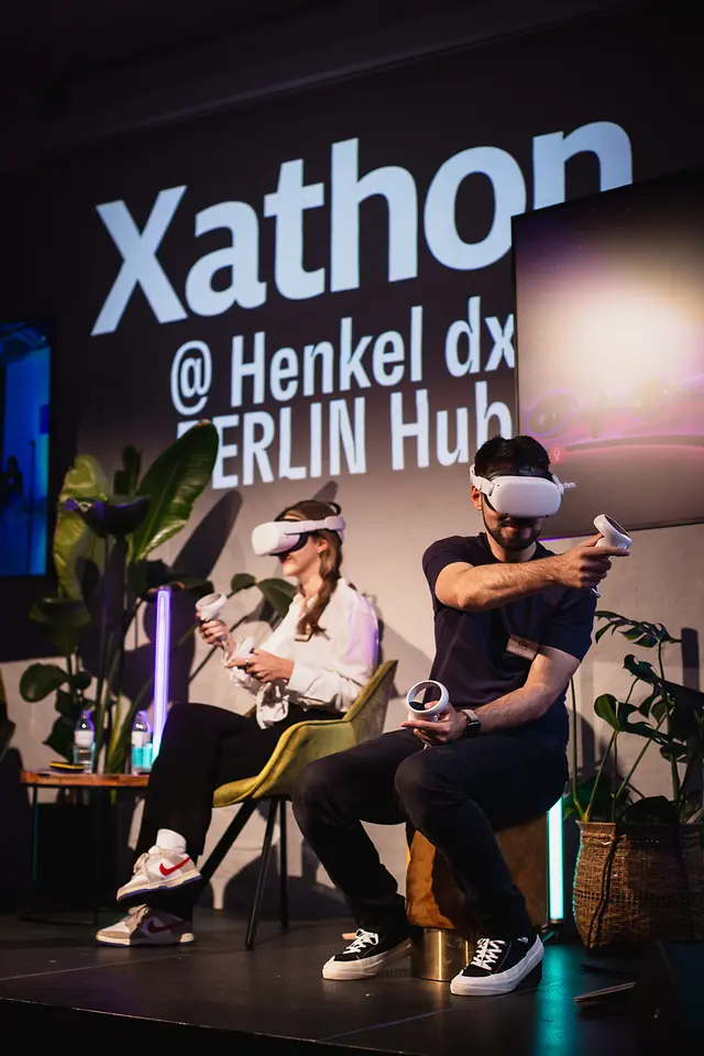 A man and a women are sitting on stage holding a remote control and wearing VR glasses.