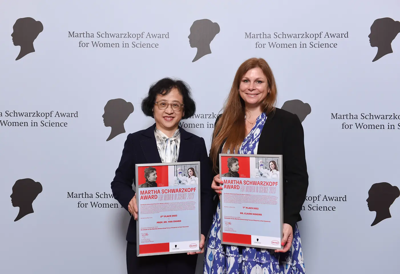 wo women standing next to each other in front of a photo wall and holding their winner's certificate. 