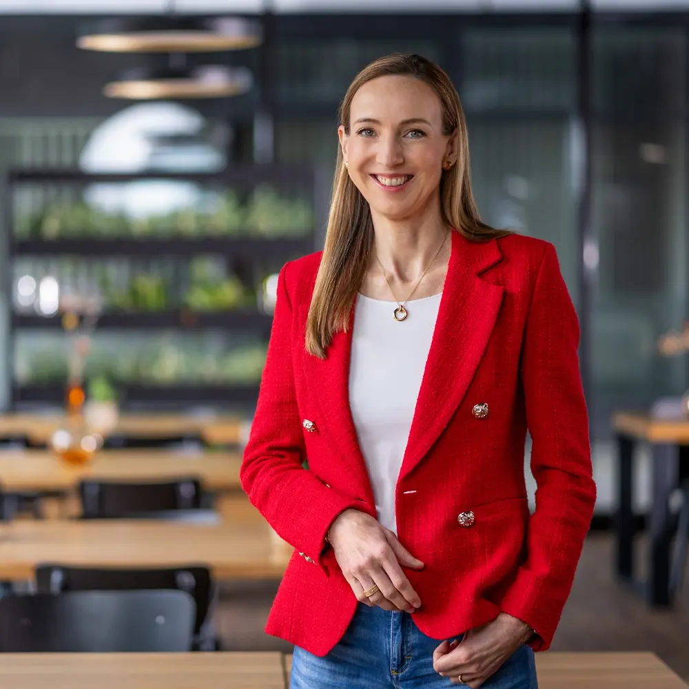 Dr. Simone Bagel-Trah v jasne červenom saku a bielom topu stojaca v modernom prostredí štýlu kaviarne, s drevenými stolmi a dekoratívnymi vázičkami v pozadí.