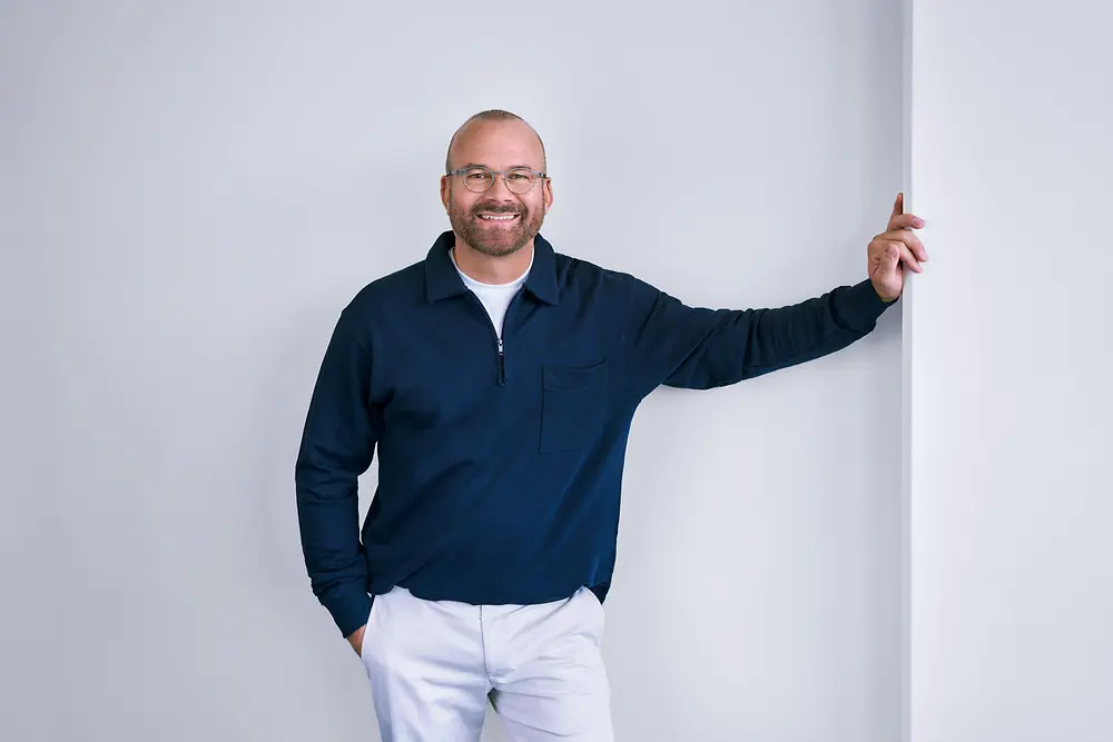 Skúsený profesionál v spoločnosti Henkel, ktorý sebavedomo stojí pri bielej stene v modernom prostredí a má na sebe tmavú košeľu s dlhým rukávom a svetlé nohavice.