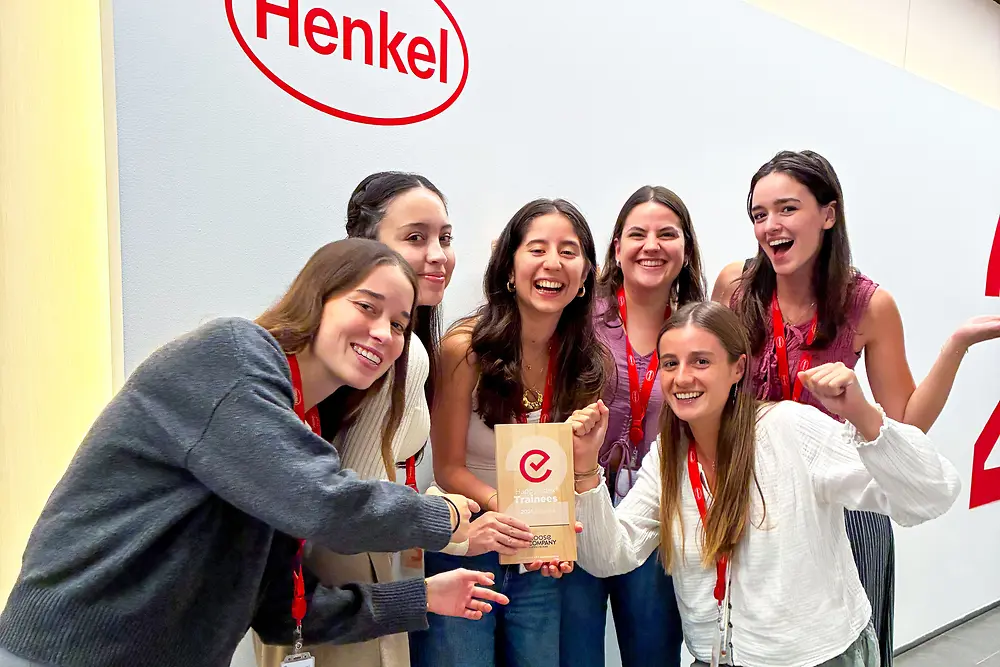 Skupina šiestich ľudí stojí spolu pred stenou s logom spoločnosti Henkel. Majú na sebe červené šnúrky a ležérne oblečenie a sú zhromaždení tesne okolo plakety s ocenením. Zdá sa, že ide o vnútorný firemný priestor so svetlou stenou a sivou podlahou. Skupina nadšene pózuje a upriamuje pozornosť na ocenenie v strede.