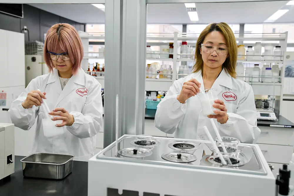 Dvaja ľudia v bielych laboratórnych plášťoch spoločnosti Henkel pracujú v modernom laboratóriu. Manipulujú s vedeckými prístrojmi a nádobami na pracovnej doske, pričom na pozadí je vidieť rôzne laboratórne vybavenie a police so zásobami.