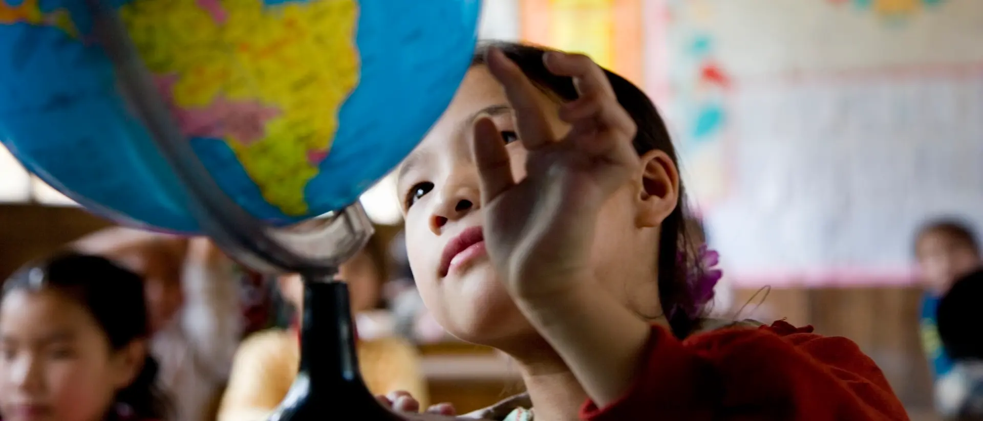 A child in a classroom reaches toward a large globe positioned on a desk, with colorful maps and posters visible on the walls in the background.