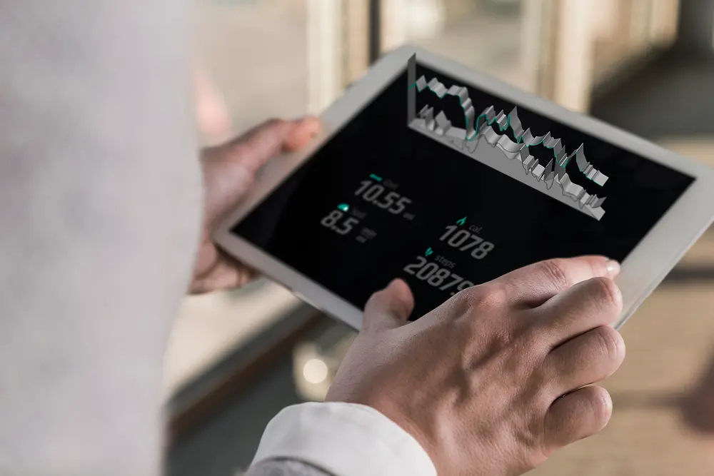 Osoba s tabletom, na ktorom sa premietajú digitálne grafy a číselné údaje, snímaná zozadu v jasne osvetlenom interiéri.