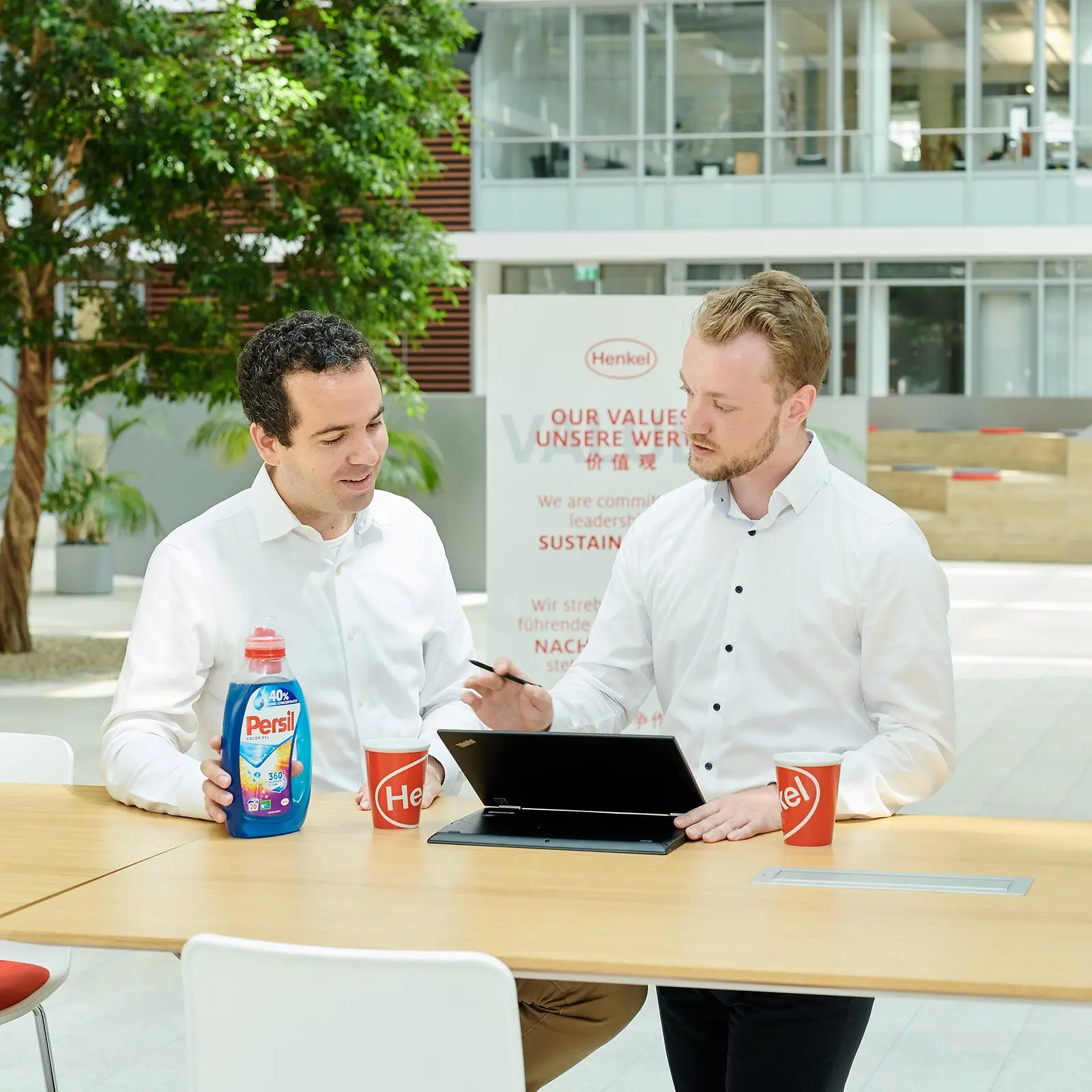 Dvaja muži stoja pri stole, na ktorom je položená nádoba Persil a laptop.